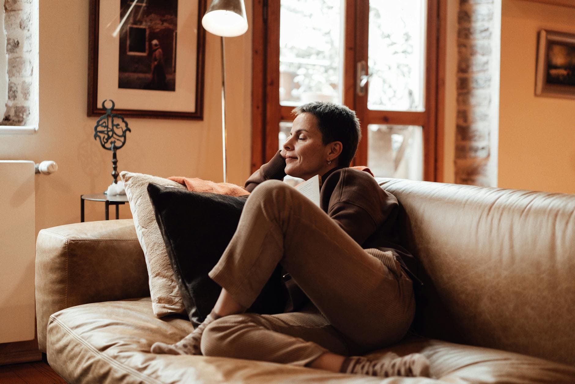 A focused, short-haired woman sitting on a comfortable couch in a sunlit room, sketching ideas on a notepad.