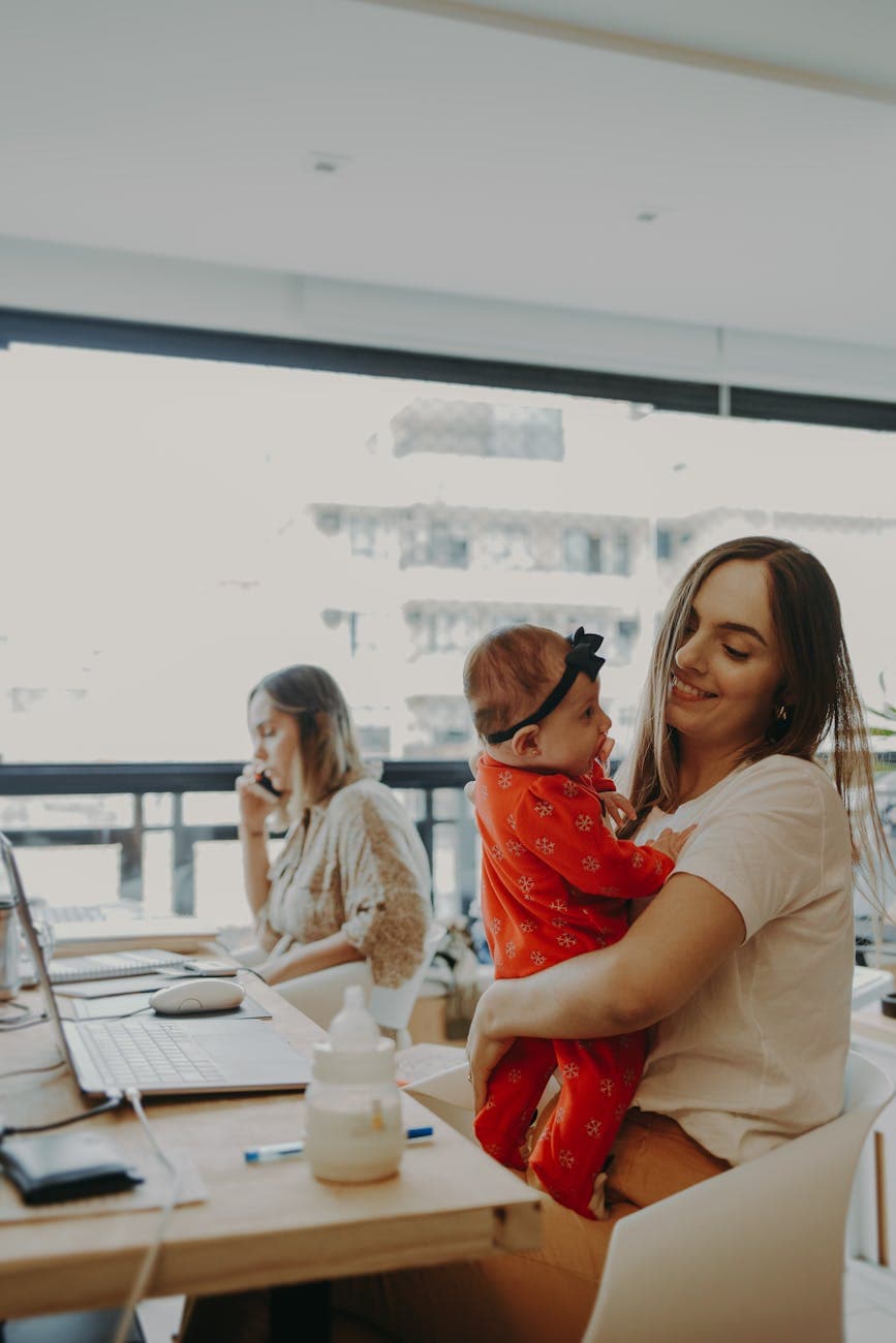 Jordan T. working on a laptop at home in a warm sunlit room while holding their toddler.